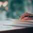 Photo of a hand with a pen and an open journal. Person is writing in a journal.