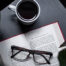 A book with a pair of classes on it with a cup of coffee next to it.