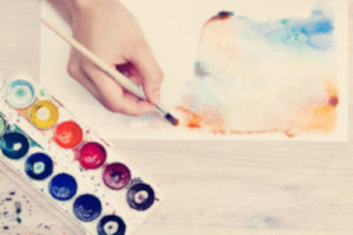 watercolorpainting-300x200-1 picture of a hand holding a paint brush and painting multicolors on a piece of paper. Watercolor paints are next to it.