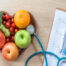 picture of fruits and vegetables in a hard shaped bowl, a stethoscope, blood glucose test and a clipboard with paper. Advertising a class on diabetes